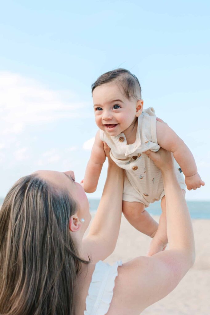 Smiling mother lifting her happy baby on the beach during a sunny day. Family portrait, candid moment, seaside photography.