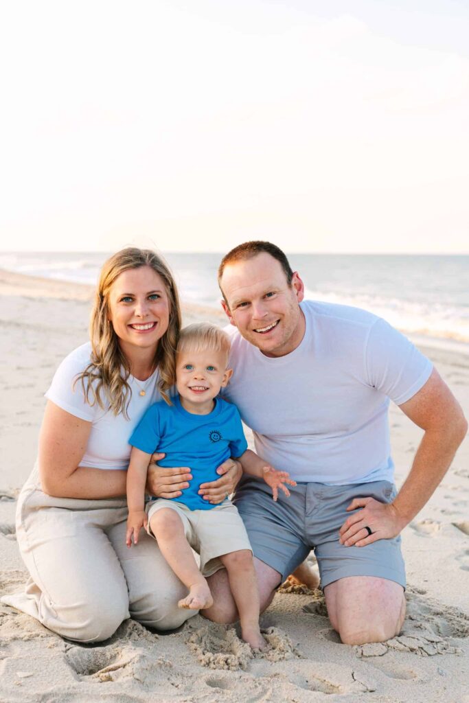Family beach portrait with smiling parents and toddler on sandy shore.
